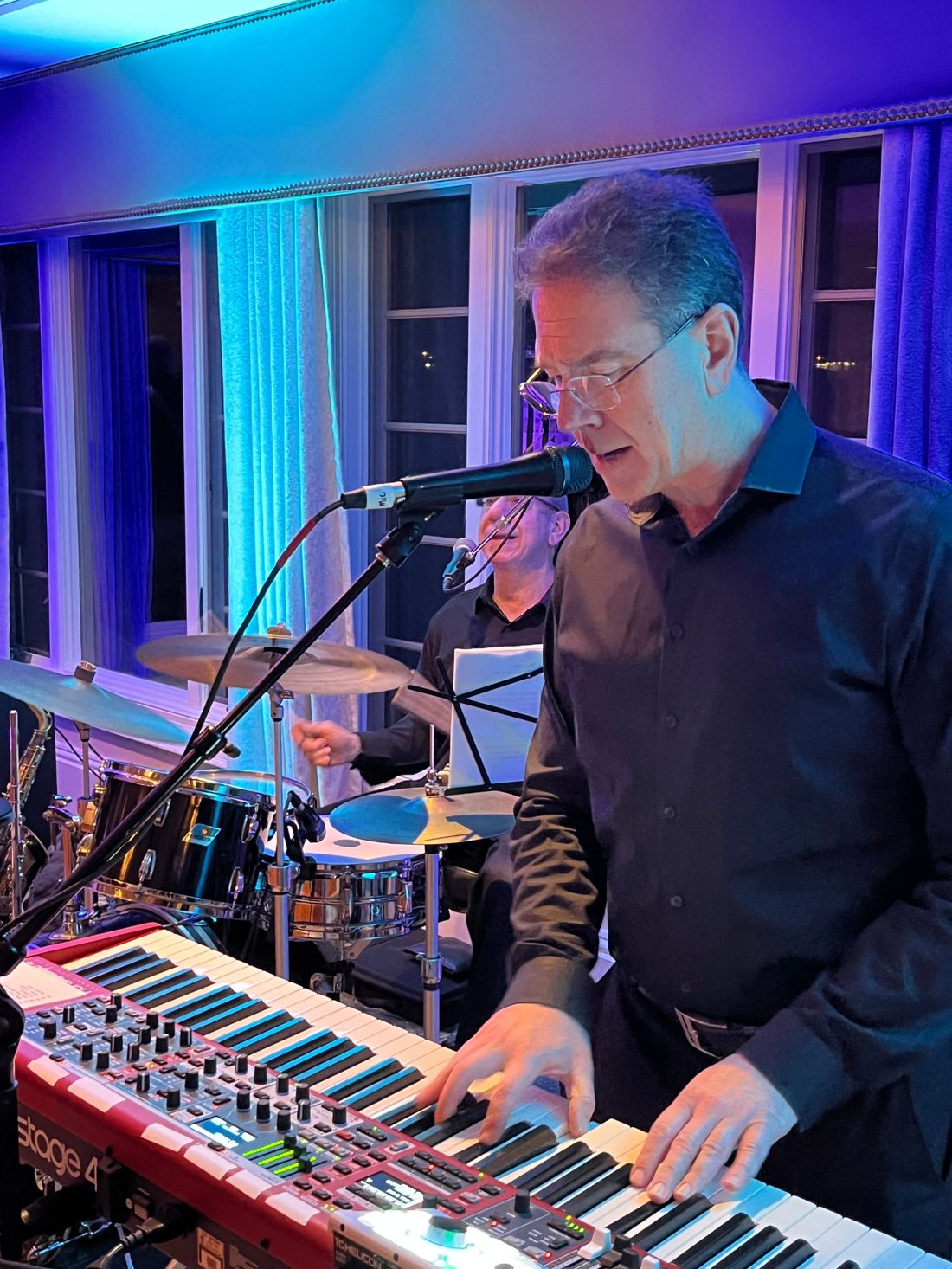 Keyboardist singing at a red Nord Stage 4 keyboard, drummer smiling behind him.